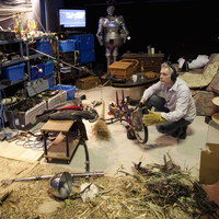 A Foley Artist seated on the floor surrounded by a variety of objects. A microphone is in front of him.